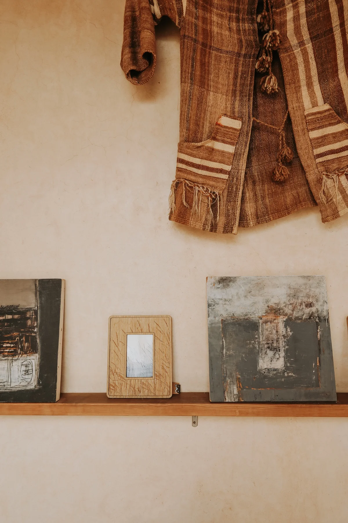 A close view of the painter's canvases on their wooden shelf, beneath a woven Berber jacket