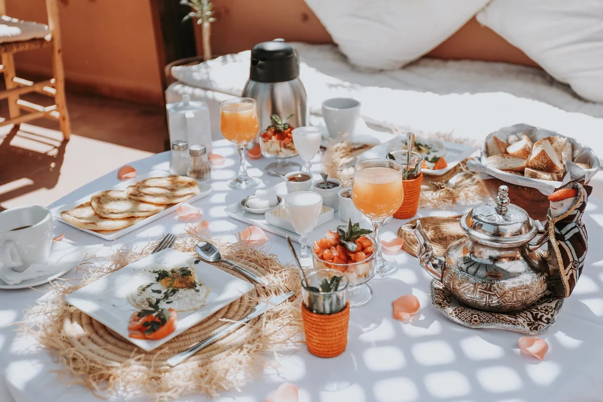 The breakfast table in dappled light — warm beghrir, eggs, pressed juices, rose petals and a silver teapot