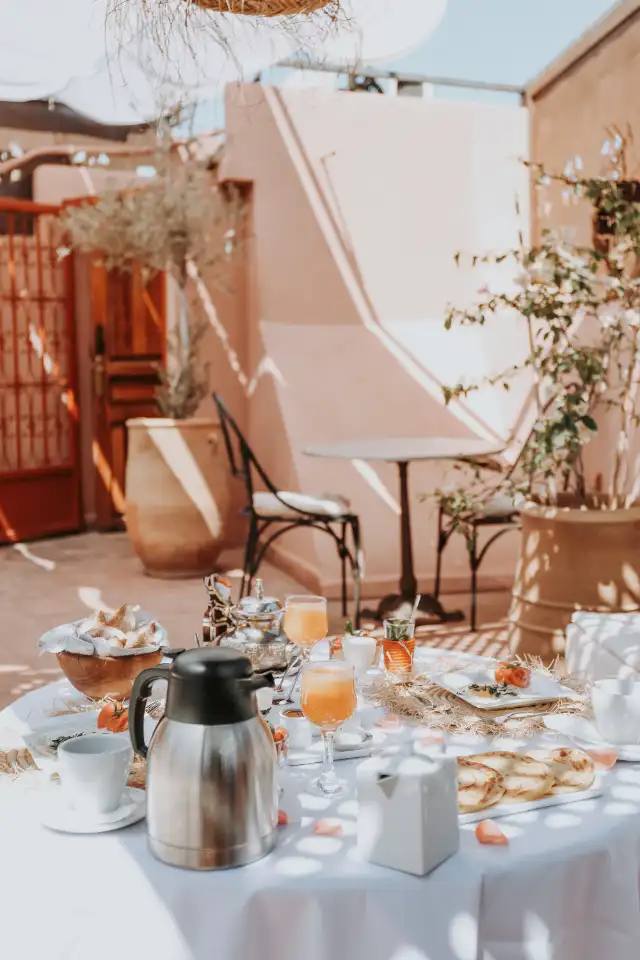 A table set on the terrace — coffee pot, orange juice, msemen, potted olive trees against pink walls